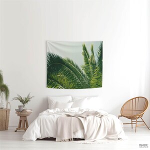 a bedroom with a large tapestry hanging on the wall depicting lush green palm leaves. The room has a white color scheme, with a bed, a chair, and a potted plant.