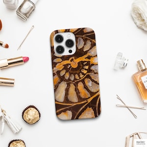 Op de afbeelding: Smartphonehoesje met een bruin, oranje en crèmekleurig fossielenpatroon. Het spiraalvormige patroon wordt benadrukt. De hoes heeft een glanzende afwerking en een uitsparing voor de camera. Schoonheidsproducten omringen de hoes.