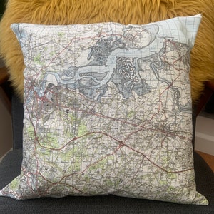 May include: A white cushion with a vintage map design printed on it. The map shows a coastal area with rivers, roads, and towns. The cushion is sitting on a fluffy yellow chair.