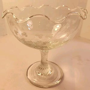 May include: Clear glass footed bowl with a scalloped rim and etched floral design. The bowl has a twisted stem and a round base. This elegant glassware is suitable for serving desserts or displaying decorative items.