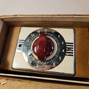 May include: Vintage timer with a cream-colored face and black numbers from 0 to 14. It has a chrome-colored metal frame and a dark red knob. The timer is in a cardboard box.