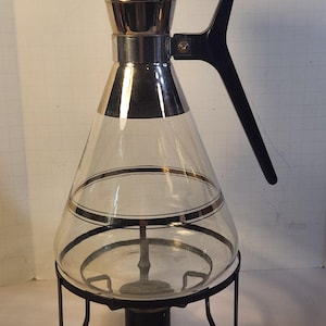 May include: A vintage glass coffee maker with a silver-toned lid and trim, and a black handle. The clear glass carafe has a black and silver band and sits on a black metal stand. A classic design for brewing coffee.