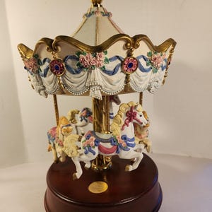 May include: A decorative carousel music box featuring white horses with pink and blue floral accents. The canopy is cream-colored with gold trim, blue ribbons, and pink roses. It stands on a circular dark wood base with a gold-colored center disc.