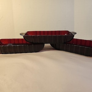 May include: Three rectangular, ruby-red glass baking dishes with ribbed sides and handles. The dishes are arranged on a light-colored surface, showcasing their deep red color and classic design. Ideal for baking and serving.