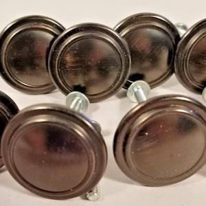 May include: Eight bronze-colored cabinet knobs with a circular design and a brushed finish. Each knob has a silver screw for attachment. The knobs are arranged in a group, suitable for furniture or home decor projects.
