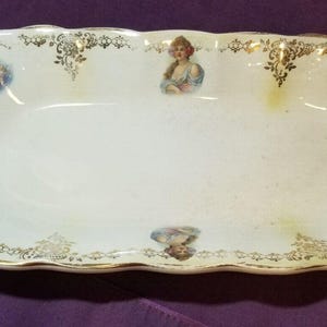 May include: An antique, oblong serving dish with a scalloped edge. The white porcelain dish features hand-painted portraits of women in blue and gold accents. Gold floral patterns adorn the rim.