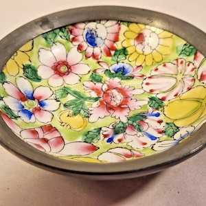 May include: A small, round dish with a dark gray metal rim. The interior is decorated with a floral pattern featuring pink, red, yellow, and blue flowers on a light green background. The dish is likely for decorative purposes.