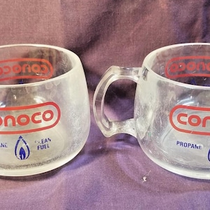 May include: Two clear glass mugs with handles. Each mug has the Conoco logo in red and white, with the words "Propane" and "Clean Fuel" and a blue flame graphic. The mugs are on a purple surface.