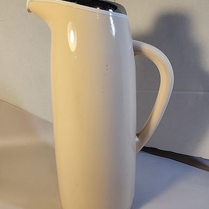 May include: A cream-colored vintage pitcher with a black and silver trim around the spout. It has a curved handle and a clear, dome-shaped lid. The pitcher is tall and cylindrical, likely for serving beverages.
