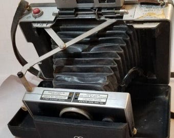 Puede incluir: Una cámara Polaroid Land vintage con una funda de cuero negro y un cuerpo plateado. La cámara tiene un flash incorporado y un visor. La cámara está en buen estado y tiene algunos arañazos menores.