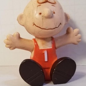 May include: A vintage figurine of a cartoon character with a round head, small ears, and a smiling face. The figure wears an orange outfit with the number 1 on the front and dark brown shoes. The arms are outstretched.