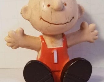 May include: A vintage figurine of a cartoon character with a round head, small ears, and a smiling face. The figure wears an orange outfit with the number 1 on the front and dark brown shoes. The arms are outstretched.