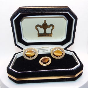 May include: A vintage jewelry set featuring cufflinks and a tie tack with tiger eye stones, presented in a black velvet box. The box has a gold trim and a gold crown logo with the words "GRAND PRIX". The cufflinks and tie tack are gold-toned.