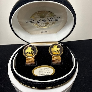May include: Gold-toned cufflinks featuring a ship design, displayed in a white and black velvet box. The box is labelled "Arts of the World" and "SWANK". The cufflinks are from Spain and are 24 carat gold.