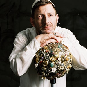 May include: A man in a white shirt and cap rests his arms on a decorative sphere. The sphere is covered in various vintage jewelry pieces, including brooches, buttons, and earrings, in gold, silver, and colorful accents. The background is a dark, textured surface.