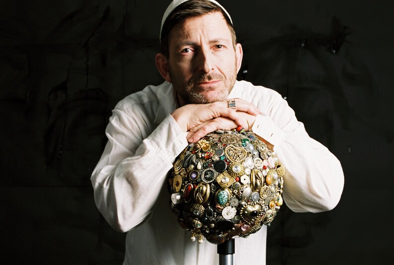 May include: A man in a white shirt and cap rests his arms on a decorative sphere. The sphere is covered in various vintage jewelry pieces, including brooches, buttons, and earrings, in gold, silver, and colorful accents. The background is a dark, textured surface.