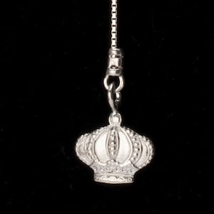 May include: A silver-colored crown pendant with intricate detailing, suspended from a delicate chain. The crown features a series of rounded arches and small, bead-like embellishments, set against a solid black background.