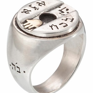 Puede incluir: Anillo de plata con una parte superior circular con símbolos grabados y una pequeña mano dorada. El anillo tiene un acabado pulido y una banda ancha. Una pieza de joyería única.