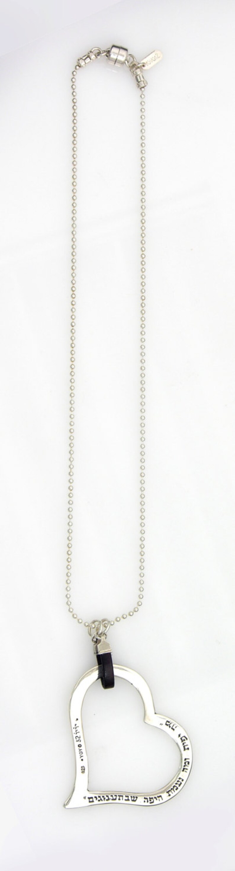 May include: A silver-toned necklace with a heart-shaped pendant. The pendant has Hebrew text and a black leather accent. The necklace chain is made of small, round beads and has a magnetic clasp.