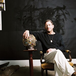 May include: A man in a black long-sleeve shirt and white pants sits in a chair, holding a decorative sphere. The sphere is adorned with various embellishments. The background features a black painted wall and various objects.