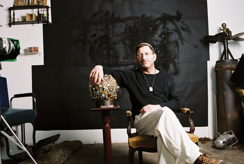 May include: A man in a black long-sleeve shirt and white pants sits in a chair, holding a decorative sphere. The sphere is adorned with various embellishments. The background features a black painted wall and various objects.
