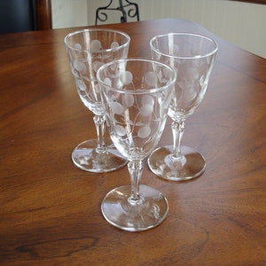 May include: Three clear glass wine glasses with an etched design of grapes and vines. The glasses are on a wooden table.
