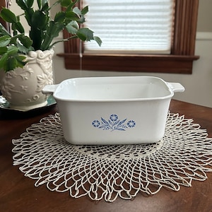May include: White ceramic baking dish with a blue floral design. The dish is sitting on a white crocheted doily on a wooden table.