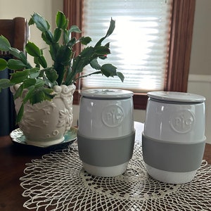May include: Two white ceramic mugs with gray silicone bands and lids. The mugs are on a white crocheted doily on a wooden table. The mugs have the word "Tile" printed on the side.
