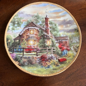 May include: A decorative plate featuring a detailed painting of a cottage scene. The artwork depicts a brick cottage with a thatched roof, surrounded by a garden with colorful flowers and a red wheelbarrow. The plate has a gold rim.