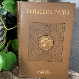 Puede incluir: Un libro antiguo de tapa dura marrón titulado "Chaucer's Poems" con letras doradas. La portada presenta un diseño ornamentado con un emblema circular central. El libro se exhibe sobre una superficie de madera.