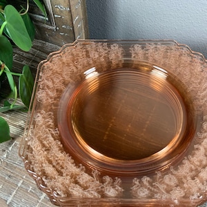 May include: A stack of six pink glass plates with a delicate floral design. The plates have a square shape with rounded corners and a scalloped edge.