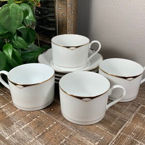 May include: Set of four white teacups with gold trim and a subtle striped pattern. The cups are arranged on a wooden surface.