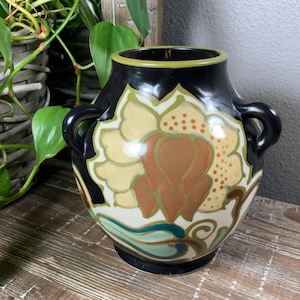 May include: A decorative ceramic vase with a black top and handles, featuring a floral design in cream, brown, and green. The vase has a black base and a yellow rim. The vase is sitting on a wooden surface.