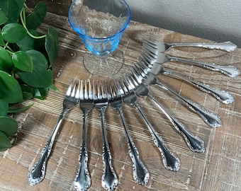Op de afbeelding: Een set van tien zilveren vorken met sierlijke handvatten, gerangschikt in een waaier op een houten oppervlak. Een blauw glas staat op de achtergrond.
