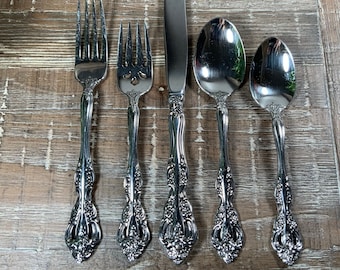 May include: Five pieces of silverware, including two forks, two spoons, and a knife, all with ornate, decorative handles. The silverware is arranged on a wooden surface.
