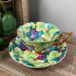 May include: A vintage teacup and saucer set with a grapevine design. The cup and saucer are decorated with green leaves, purple grapes, and gold accents.