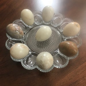 May include: A glass serving tray with eight smooth, white and brown eggs arranged in a circle. The tray has a decorative, scalloped edge and a textured, raised pattern.
