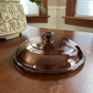 May include: A brown glass lid with a rounded shape and a small, dark brown knob. The lid is transparent, allowing light to pass through, and sits on a wooden surface. The lid is likely for a casserole dish or similar cookware.