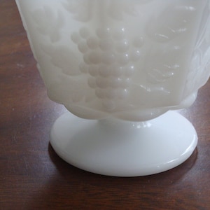 May include: White milk glass vase with a grape design. The vase has a pedestal base.