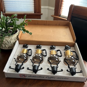 May include: Four glass coffee makers with black handles and bases, gold accents, and black tops are arranged in a cardboard box. The box has the text "For distinctive Coffee service" printed on it.