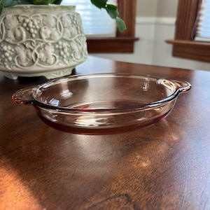 May include: A clear, round, glass baking dish with two small handles. The dish has a reddish-brown tint and is empty. A decorative, light green, ceramic pot with a plant is in the background.
