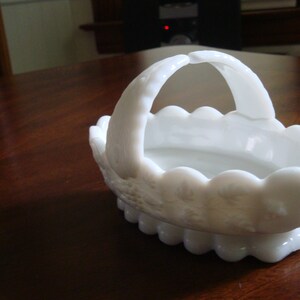 May include: White milk glass basket with a scalloped edge and a handle. The basket is decorated with a floral pattern.