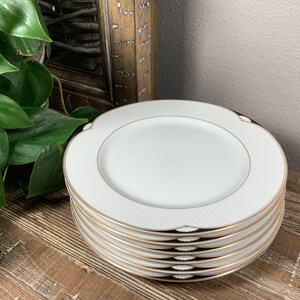 May include: A stack of six white dinner plates with a gold rim and a subtle embossed pattern. The plates are stacked on a wooden surface.