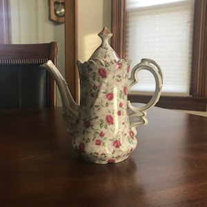 May include: A white porcelain teapot with a floral pattern of pink roses. The teapot has a gold trim and a handle with a decorative design.