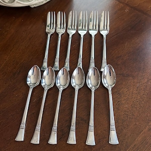 May include: A set of vintage silverware, including forks and spoons, arranged on a dark wooden surface. The silverware has a shiny, silver finish with decorative detailing on the handles. The spoons and forks are arranged in parallel rows.
