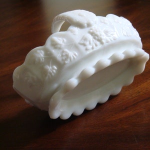 May include: White milk glass bowl with a scalloped edge and a raised floral design. The bowl has a handle and is in the shape of a basket.