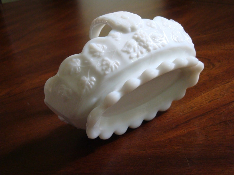 May include: White milk glass bowl with a scalloped edge and a raised floral design. The bowl has a handle and is in the shape of a basket.
