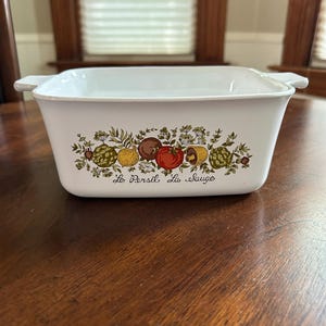 May include: White ceramic baking dish with a floral design featuring green herbs, red tomatoes, yellow lemons, and brown mushrooms. The dish has two handles and the text "Le Persil Le Sauge" on the bottom.