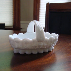 May include: White milk glass basket with a scalloped edge and a raised floral design. The basket has a handle and is perfect for serving snacks or holding small items.