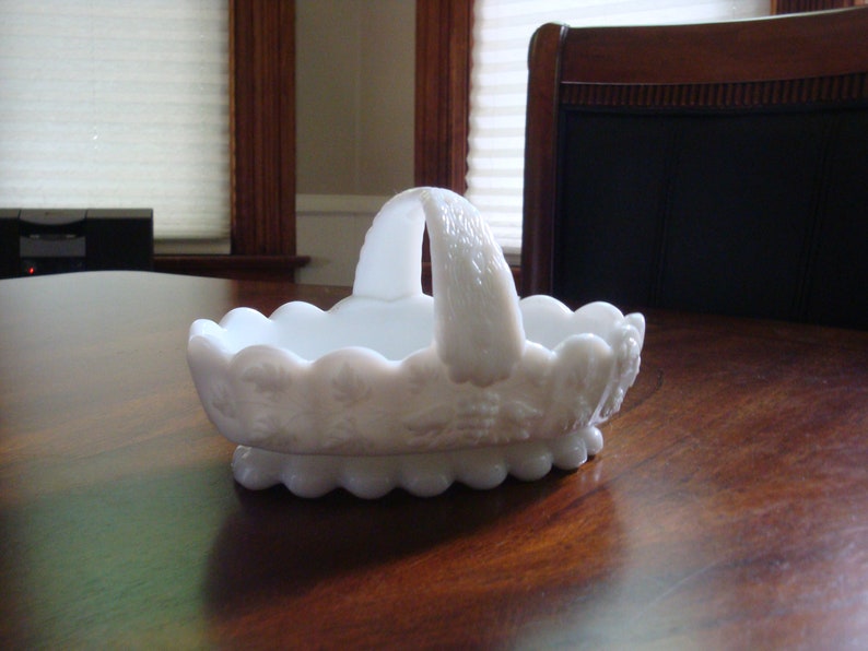 May include: White milk glass basket with a scalloped edge and a raised floral design. The basket has a handle and is perfect for serving snacks or holding small items.
